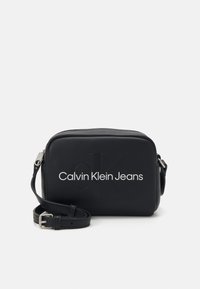 Borsa a tracolla nera in pelle con tracolla regolabile, logo Calvin Klein Jeans impresso, forma rettangolare e superficie liscia.