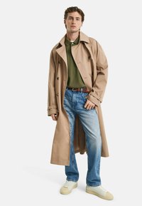 Beige trenchcoat med dubbelknäppt design, grön polo-tröja, ljusblå jeans och vita sneakers med beiga accenter.