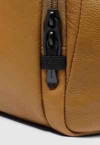 Hoek van een leren tas in tan kleur met een zwarte ritssluiting, een gestructureerde afwerking en een geweven banddetail. Voorzien van een duurzame rits voor gemakkelijke toegang.