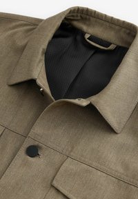 Olive green jacket with a fold-down collar, textured fabric, black mesh lining, and a black button detail at the front.