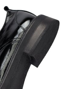 Zwarte gepolijste leren schoen met een gestructureerde zwarte rubberen zool, voorzien van een ronde neus en een lage hak. Stiksels langs de bovenkant.