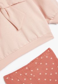 Sudadera rosa empolvada con detalle de lazo y dobladillo acanalado, combinada con pantalones cortos coral con patrones de corazones color crema. Suave mezcla de algodón ligero.