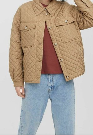 Person wearing a tan quilted jacket with buttoned flap pockets, a maroon shirt, and light blue high-waisted jeans against a plain background.
