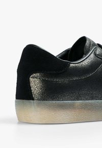 Zapatilla negra de corte bajo con suela de beige texturizada, que cuenta con un acabado metálico suave en el costado y una pestaña de talón de ante negro suave.