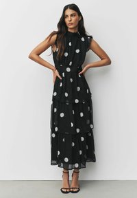 Schwarzes ärmelloses Kleid mit großen weißen Punkten, Rüschenausschnitt und mehrlagigem Stoffdesign. Kombiniert mit schwarzen Riemchenheels.