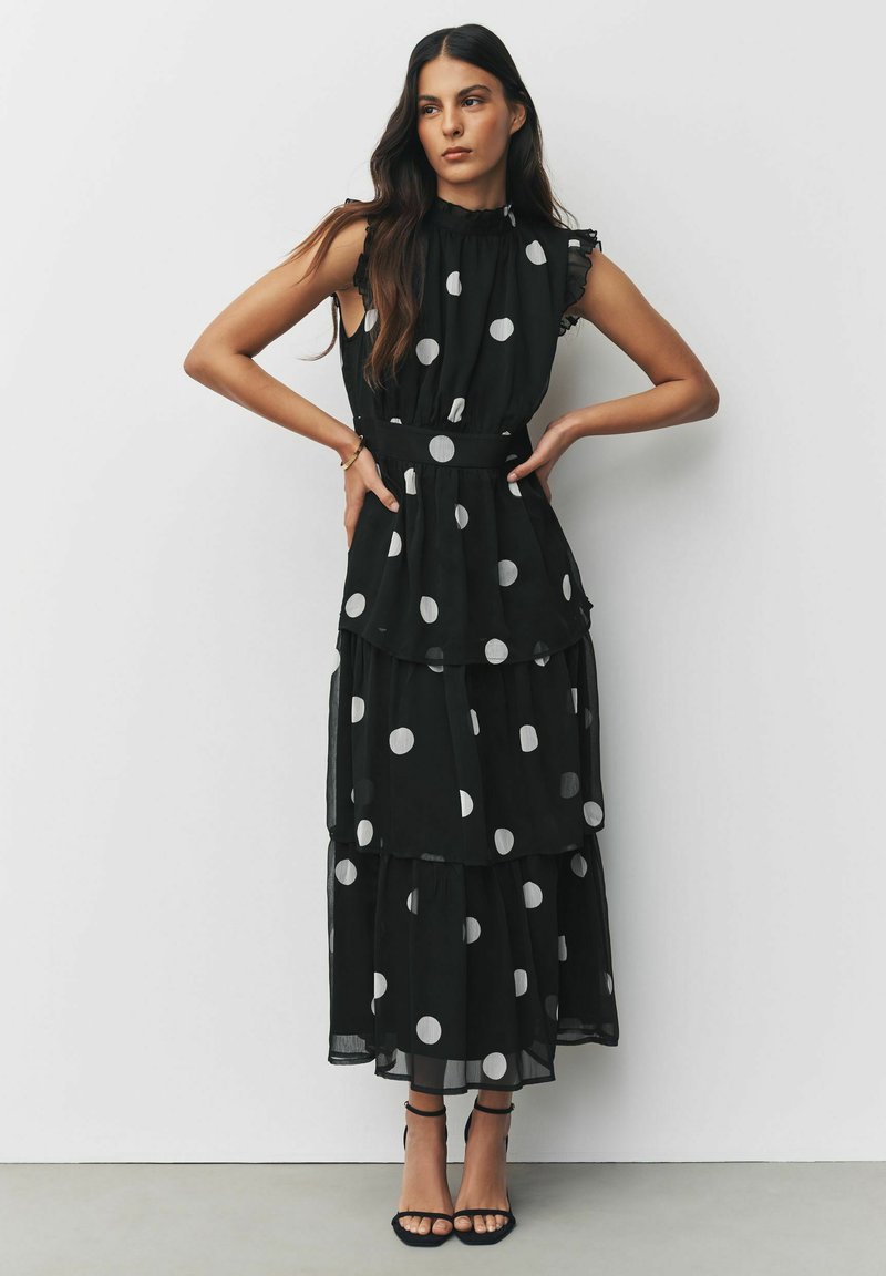 Schwarzes ärmelloses Kleid mit großen weißen Punkten, Rüschenausschnitt und mehrlagigem Stoffdesign. Kombiniert mit schwarzen Riemchenheels.