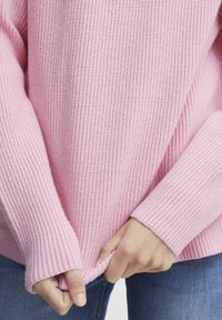 Svetlo roza rebrast pulover z dolgimi rokavi, z sproščenim krojem in strukturirano površino, oblečen čez modre kavbojke.