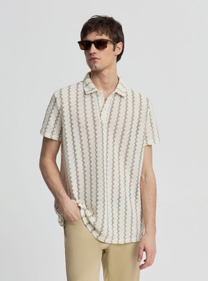 Giovane uomo con occhiali da sole marroni, camicia beige a maniche corte con motivo e pantaloni marrone chiaro, in piedi con una mano in tasca.