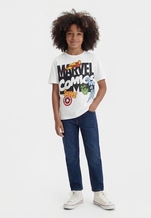 Barn med krøllet hår iført en hvid Marvel Comics T-shirt, blå jeans og hvide sneakers, står med den ene hånd i lommen.