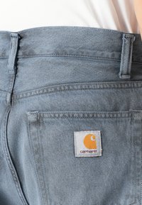 Lyseblå denimbukser med rett snitt, som har en baklomme og et fremtredende oransje Carhartt-logo merket på hvit bakgrunn.