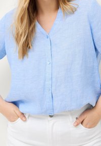 Camicia azzurro chiaro a maniche corte, con bottoni, realizzata in un tessuto testurizzato. Presenta un colletto a V e una leggera lucentezza, abbinata a pantaloni bianchi.