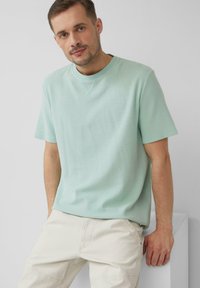 Jasnozielona bawełniana koszulka z okrągłym dekoltem i krótkimi rękawami, z fakturowanym materiałem i widocznymi detalami szwów. Połączona z jasnobeżowymi spodniami.