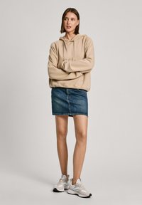 Sudadera beige con capucha y un corte relajado, combinada con una falda mini de mezclilla y zapatillas gris claro. Diseño sencillo, con una textura suave.
