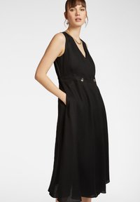 Abito nero senza maniche con scollatura a V, silhouette a trapezio, dettagli in vita con bottoni argentati e tasche laterali. Tessuto morbido e leggero.