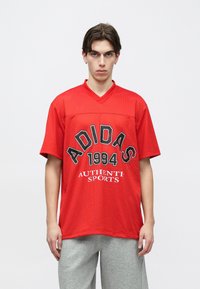 Νέος άνδρας με κόκκινη αθλητική φανέλα Adidas με το κείμενο «1994 Authentic Sports» και γκρίζο φούτερ παντελόνι, στέκεται μπροστά σε απλό λευκό φόντο.