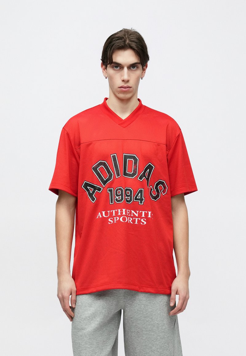 Jeune homme portant un maillot de sport rouge Adidas avec le texte « 1994 Authentic Sports » et un pantalon de survêtement gris, debout devant un fond blanc uni.
