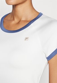 Hvit atletisk topp med korte ermer, med en mørkeblå krage og ermetrim. Inkluderer en liten gylden logo på venstre bryst.
