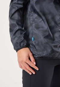 Giacca nera leggera con finitura lucida, polsini elastici e etichetta laterale con logo in blu e giallo. Indossata sopra leggings neri.