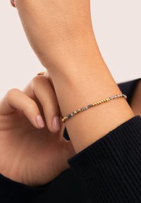 Armband med guld- och grå kulor som har växelvis polerade guldkulor och strukturerade grå kulor, bärs på ett handled mot en mörk tröja.