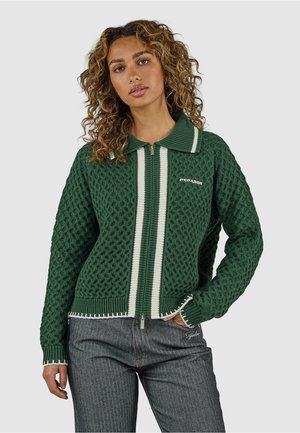 Kvinde med krøllet hår iført en grøn tekstureret lynlåssweater med hvide striber og mørkegrå bukser, stående mod en ensfarvet baggrund.