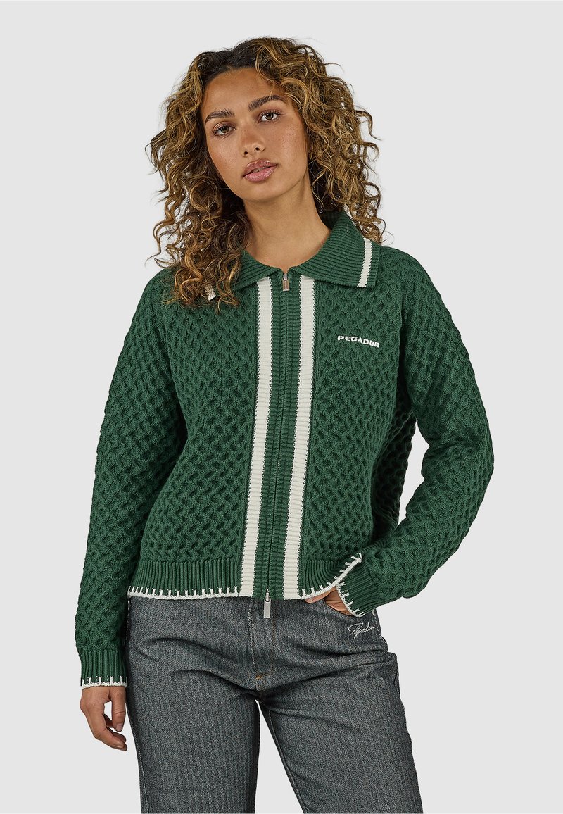 Žena s kučeravými vlasmi v zelenej textúrovanej mikine so zapínaním na zips, bielymi pruhmi a tmavosivých nohaviciach, stojaca pred jednoduchým pozadím.