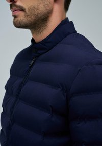 Mann mit leichtem Bart, der eine dunkelblaue gepolsterte Jacke mit Reißverschluss und hohem Kragen vor einem schlichten Hintergrund trägt.