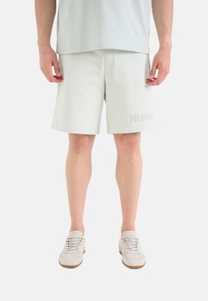 Hombre vistiendo pantalones cortos beige claro con "HUGO" grabado en la pierna izquierda, combinados con zapatillas claras a juego, de pie contra fondo blanco.