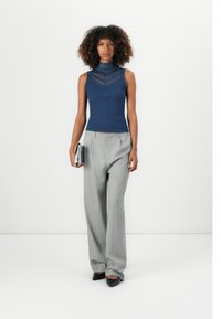 Mulher com cabelo encaracolado, vestindo uma blusa sem mangas em renda azul-marinho e calças largas cinzas, segurando uma carteira prateada, de pé contra um fundo branco.