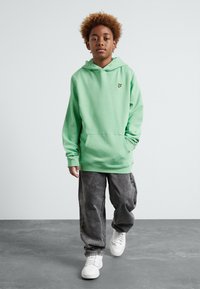 Sudadera verde clara con bolsillo canguro y pequeño logo amarillo en el pecho, combinada con pantalones vaqueros grises holgados y zapatillas blancas.