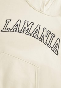 Krém színű kapucnis pulóver fekete "LAMANIA" felirattal ívelt formában. Elöl kenguruzseb található, és puha, texturált anyagból készült.