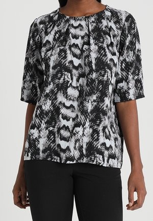 Blouse à manches courtes à motif abstrait noir et blanc portée avec un pantalon noir, visible des épaules aux hanches.