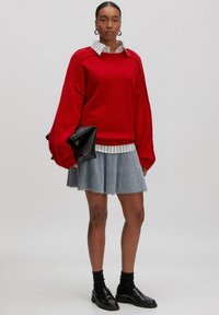 Vrouw in een rode oversized trui over een gestreept shirt, een grijze fuzzy rok, zwarte sokken en loafers, met een gevouwen zwarte clutch in haar hand.