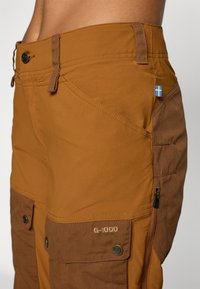 Bruine outdoor broek met meerdere zakken, knoop- en ritsdetails, G-1000 logo en een klein Zweeds vlaggetje aan de zijkant.