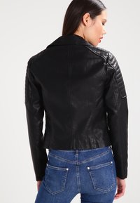 Black leather jacket with quilted shoulder accents, fitted design, and a cropped length. Paired with blue jeans, showing rear view.