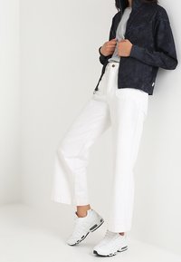 Námořnická zipová bunda s texturovaným vzorem, v kombinaci s bílými kalhotami se širokými nohavicemi a bílými teniskami s šedými akcenty.