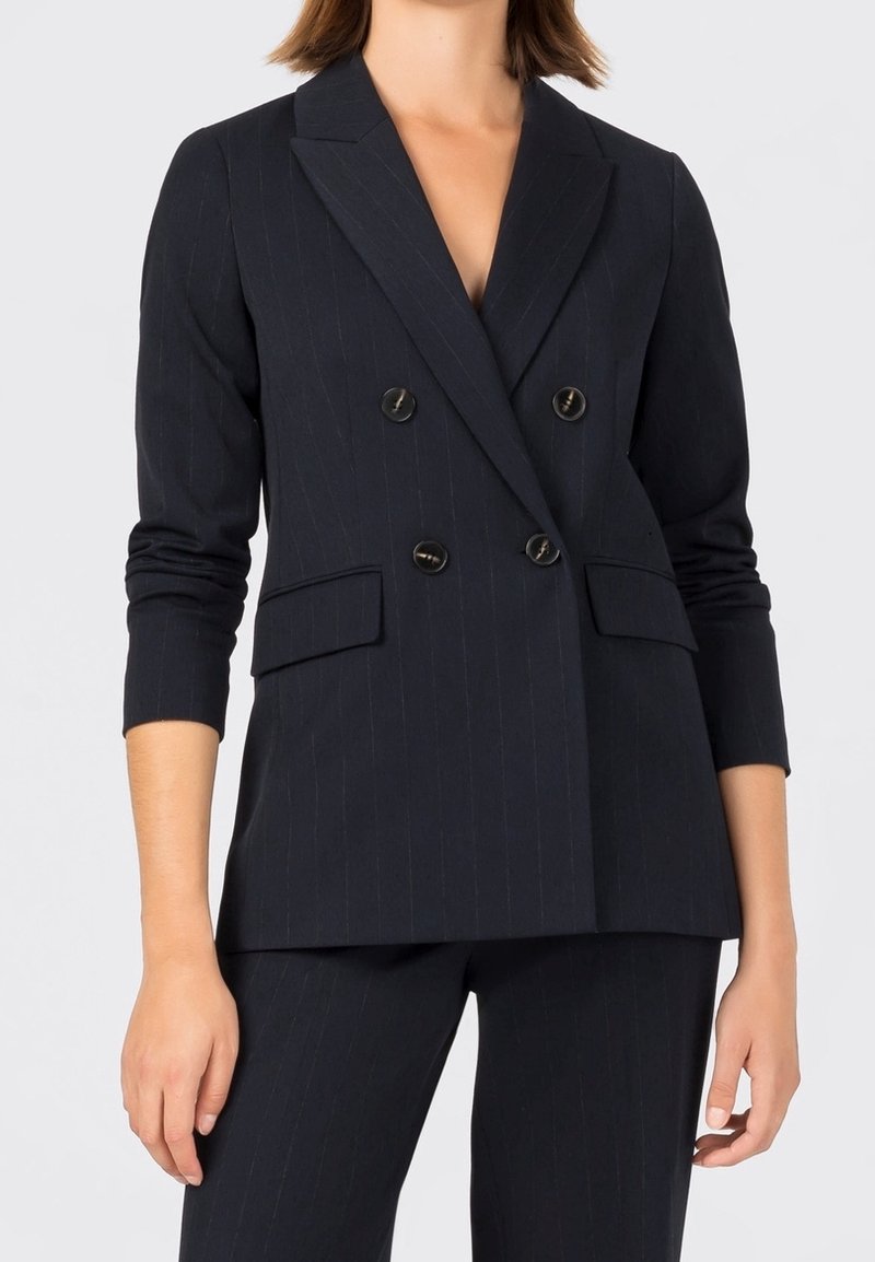 Femme portant un blazer croisé noir à fines rayures avec quatre boutons et un pantalon assorti, debout sur un fond uni.