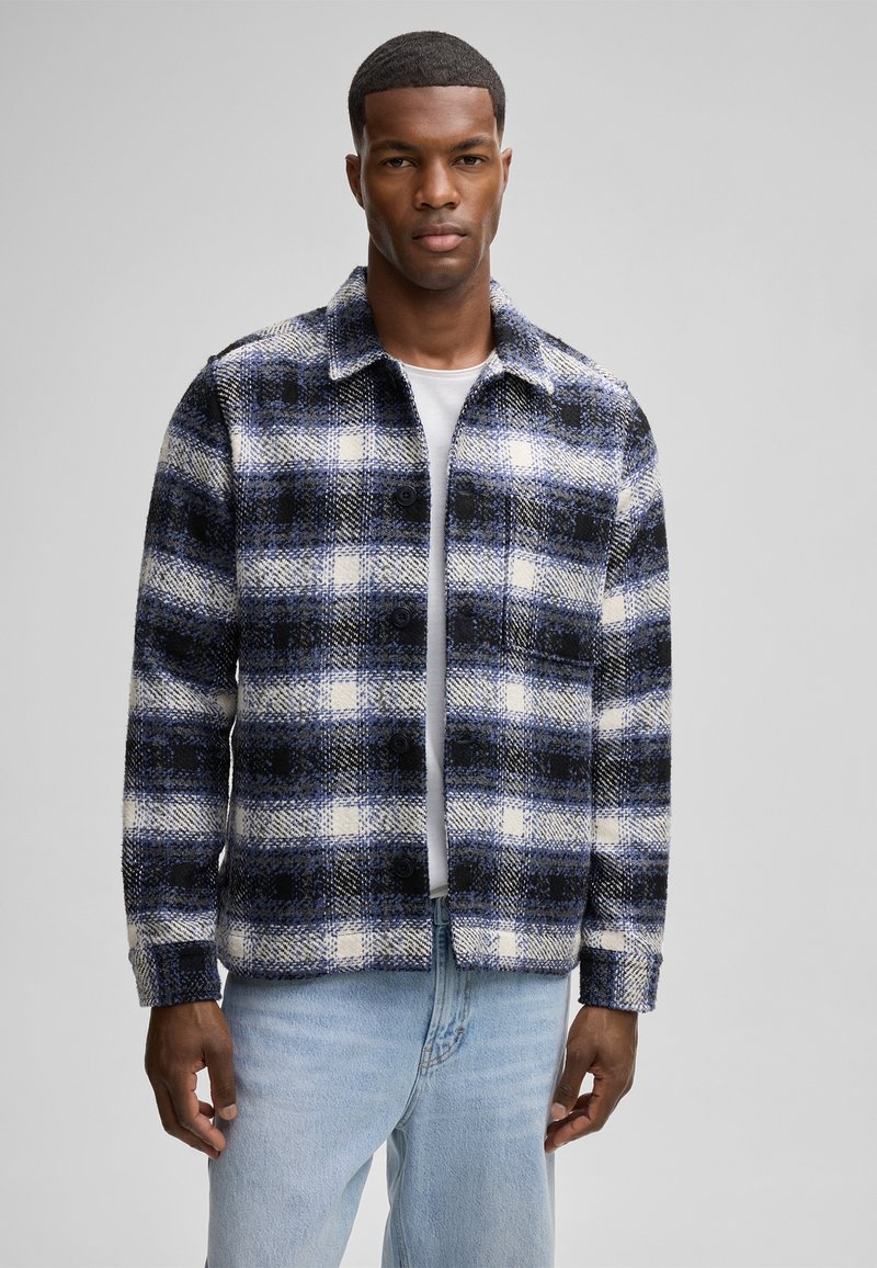 Kariertes Jackett in Blau, Schwarz und Weiß; mit Kragen, Knopfleiste und langen Ärmeln, kombiniert mit hellblauen Jeans.