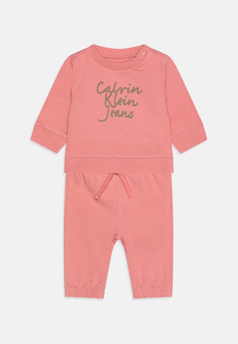 Svetloružový dvojdielny outfit pozostávajúci z topu s dlhým rukávom so strieborným nápisom "Calvin Klein Jeans" a zodpovedajúcich teplákov s elastickými manžetami.