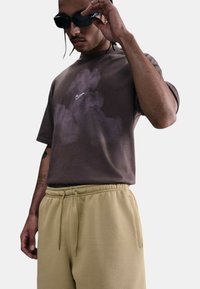 Camiseta marrón con un logo de Nike blanco y un patrón de nubes, combinada con pantalones joggers beige que tienen una cintura elástica y bolsillos laterales.