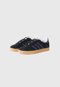 Juodos suede sportbačiai su violetiniais trimis juostelėmis, balta liežuvėlio dalimi ir guminiu padu. Suapvalinta nosis ir plokščios raištelių varstymo detalės.