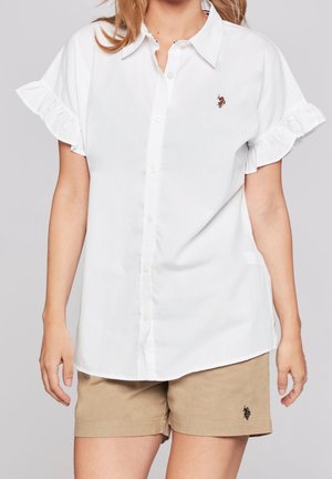 Vrouw draagt een witte korte-mouwen blouse met knopen en ruches aan de mouwen en beige shorts, beide met kleine geborduurde logo's.