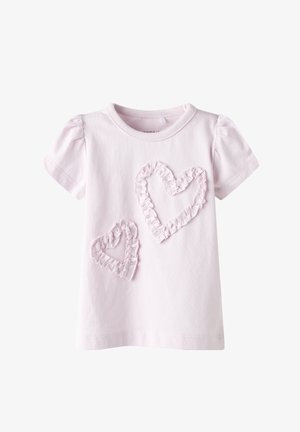 Camiseta de algodón rosa claro con mangas cortas abullonadas y dos aplicaciones en forma de corazón con volantes en un rosa ligeramente más oscuro. Textura suave.