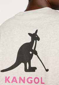 Világosszürke pamut póló, nagy fekete kenguru grafikával, amely golfütőt tart, az alján rózsaszín "KANGOL" felirattal.