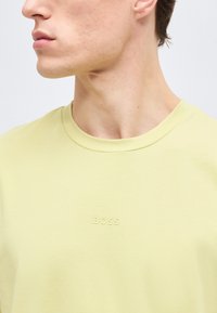 Άνδρας που φοράει ανοιχτό κίτρινο T-shirt με λαιμόκοψη και ανάγλυφο λογότυπο "BOSS" στο στήθος, στραμμένος προς τα αριστερά με ορατή γνάθο και λαιμό.