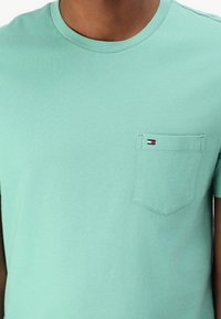 Light green cotton t-shirt featuring a round neckline, short sleeves, and a left chest pocket with a contrasting logo patch.