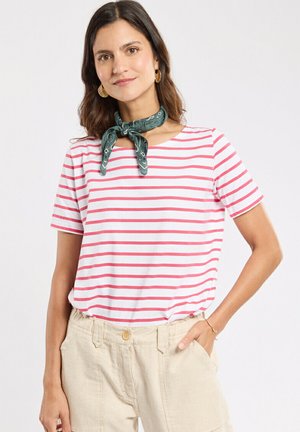 Femme portant un haut à manches courtes rayé blanc et rose, un pantalon beige taille haute, un foulard vert à motifs autour du cou et des boucles d'oreilles en or, avec la main dans la poche.
