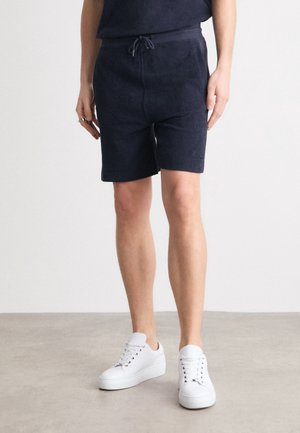 Person trägt dunkelblaue Shorts mit Kordelzug und weiße Schnürsneaker, steht auf einem hellgrauen Boden vor einer weißen Wand.