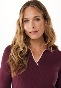 Pletený pulovr v burgundské barvě s žebrovanou strukturou, s výstřihem do V obšitým světle růžovým lemem. Dlouhé vlnité vlasy částečně viditelné.