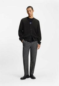 Svart stickad cardigan med V-ringning och fem knappar, i kombination med grå texturerade byxor. Modellen bär svarta skor och står framför en vit bakgrund.