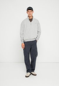 Sudadera de felpa gris con cremallera y puños de canalé, combinada con pantalones cargo navy que tienen bolsillos laterales y detalles en gris. El modelo lleva zapatillas claras.